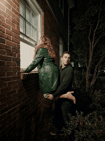 Marcie in a green leather coat is hoisted up to peer through a brick-building window at night while Ben, holding her up, glances nervously over his shoulder.