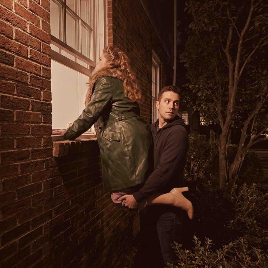 Album-cover crop of the show photo: Marcie in a green leather coat is hoisted up to peer through a brick-building window at night while Ben, holding her up, glances nervously over his shoulder.